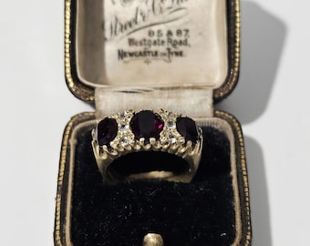 Puede incluir: Un anillo de oro con tres piedras de color rojo oscuro de corte ovalado y pequeñas piedras transparentes, presentado en un joyero vintage de cuero marrón con forro color crema y el texto Street & Co Ltd, 85 & 87, Westgate Road, Newcastle on Tyne.