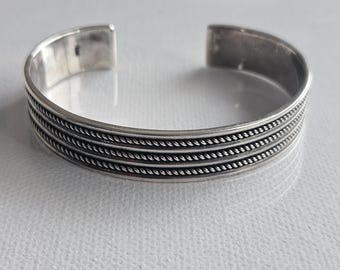 Pode incluir: Uma pulseira tipo bracelete de prata com acabamento polido e três filas horizontais de detalhes em corda trançada ao longo do centro. O design aberto apresenta uma superfície interna lisa e um exterior arredondado ligeiramente oxidado.