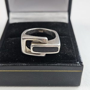 May include: A silver ring featuring an interlocking geometric design with a smooth, rectangular black inlay. The ring has a polished metallic finish and is presented inside a black jewellery box with a soft, dark interior lining.