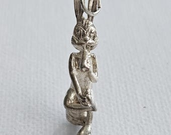 Pode incluir: Um pingente de metal prateado em forma de coelho de desenho animado sentado enquanto segura uma pequena trombeta na boca. O coelho tem orelhas longas e eretas e traços faciais detalhados, com um anel circular preso ao topo da cabeça.