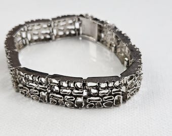 Pode incluir: Uma pulseira de elos de metal prateado com um padrão vazado repetitivo de segmentos retangulares. O design consiste em duas fileiras horizontais de aberturas texturizadas tipo grade conectadas por dobradiças e um fecho de metal seguro.