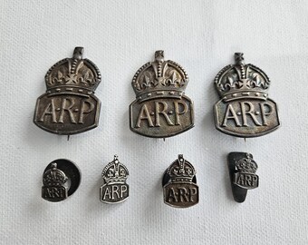 Pode incluir: Uma coleção de sete emblemas e alfinetes de metal prateado ARP, cada um apresentando um emblema de coroa acima das letras ARP. O conjunto inclui três emblemas maiores em forma de escudo e quatro alfinetes menores de várias formas metálicas.