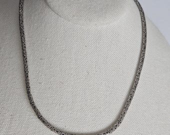 Pode incluir: Um colar de tom prateado com um padrão contínuo de elos de corrente bizantina. A corrente de metal tem uma aparência arredondada e texturizada com segmentos interligados que criam um design uniforme e flexível ao longo de toda a peça.