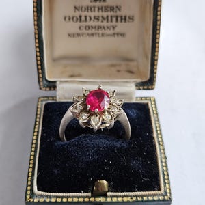 May include: A silver-toned ring featuring a central oval-cut vibrant red gemstone surrounded by a halo of small clear stones. The ring is presented in a dark green velvet-lined jewelry box with the text The Northern Goldsmiths Company Newcastle-on-Tyne.