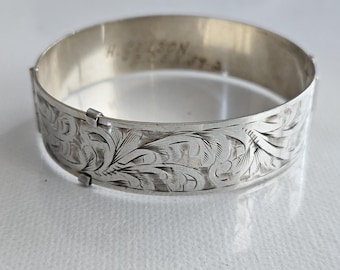 Pode incluir: Um bracelete de metal cor de prata com um intrincado padrão floral gravado na sua superfície exterior. O interior da banda apresenta uma inscrição ténue que diz H. NELSON, com um mecanismo de dobradiça visível na lateral.