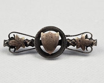 Pode incluir: Um broche de barra horizontal com uma moldura circular central contendo um pequeno emblema em forma de escudo. O broche é feito de metal oxidado escuro e acentuado com dois motivos de folhas em tom dourado em cada lado do círculo central.