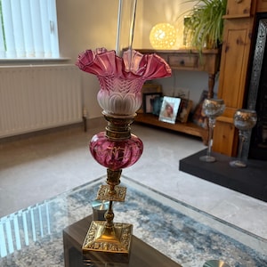 Op de afbeelding: Een olielamp met een vierkante koperen voet met gedetailleerde versieringen, een centraal bolvormig magenta glazen reservoir en een geplooide magenta en witte glazen kap. Een hoge, heldere glazen cilinder steekt uit de bovenkant van de lamp.