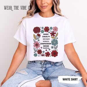 Puede incluir: Una camiseta blanca de manga corta con un gráfico central de flores coloridas que rodean el texto en negro: WHEN CRUELTY BECOMES NORMAL COMPASSION SEEMS RADICAL. El diseño floral incluye tonos rojos, rosas, azules y verdes.