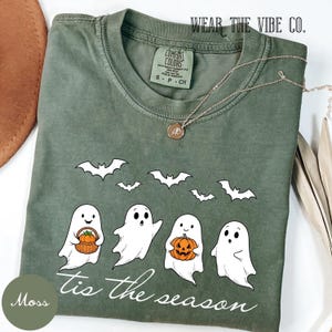 Puede incluir: Una camiseta de manga corta color verde musgo con un gráfico de cuatro fantasmas blancos, cinco murciélagos negros y el texto 'tis the season' en letra blanca. Un fantasma sostiene una cesta marrón con calabaza y otro una calabaza naranja tallada.