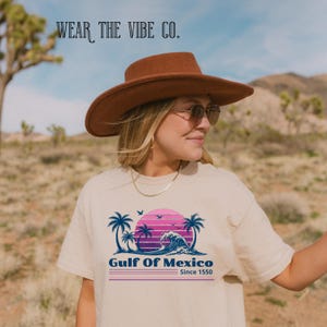 Puede incluir: Una camiseta beige de manga corta con un estampado gráfico de palmeras, una ola y una puesta de sol en tonos rosa y morado. El texto dice "Gulf Of Mexico Since 1550" y "WEAR THE VIBE CO.". Se lleva con un sombrero marrón de ala ancha.