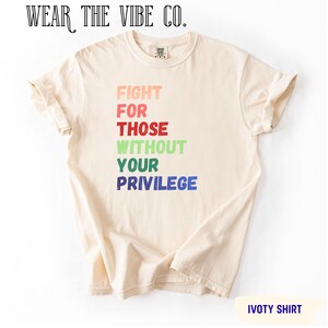 Può includere: Una t-shirt a maniche corte color crema con il testo "FIGHT FOR THOSE WITHOUT YOUR PRIVILEGE" stampato in righe sovrapposte di vari colori, tra cui pesca, rosa, rosso, verde chiaro, verde scuro e blu. Il marchio "WEAR THE VIBE CO." è riportato sopra.