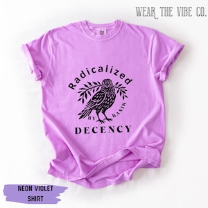 May include: A neon violet short-sleeve t-shirt featuring a black graphic of a bird holding a branch. The text reads Radicalized by Basik Decency. The shirt has a crew neckline and a relaxed fit, with the brand name Wear The Vibe Co. visible.