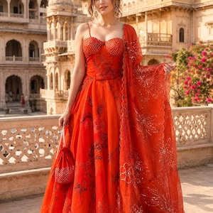 Könnte beinhalten: Ein leuchtend oranges bodenlanges Kleid mit einem strukturierten Korsett-Oberteil mit dünnen Trägern und einem weiten Rock mit dunklem Blumenmuster. Das Set enthält einen passenden transparenten orangen Dupatta mit Paillettenstickerei und eine kleine Tasche mit Perlenhenkel.