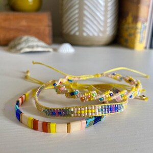 Può includere: Tre braccialetti fatti a mano con perline di vetro colorate. Un braccialetto ha perline rettangolari piatte gialle, arancioni, rosa e blu. Un altro è una fascia intrecciata con un motivo geometrico. Il terzo è un cordino annodato con piccole perline.