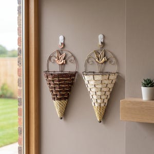 May include: Two wall-mounted cone-shaped baskets featuring a metal frame with a bird ornament at the top. One basket is woven with dark brown material, while the other is woven with cream-colored material. Both have a yellow and white striped tip.