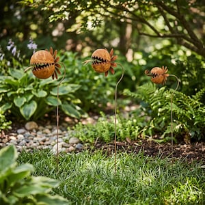 May include: Three garden stakes featuring spherical, rusted metal plant heads with jagged, tooth-like openings and protruding tongues. Each sculpture is mounted on a thin, vertical metal rod, varying in size from largest to smallest.
