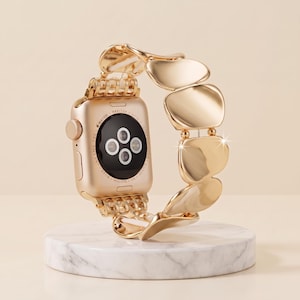 Pode incluir: Um smartwatch de tom dourado com uma pulseira de elos metálicos composta por segmentos lisos, irregulares e em forma de seixo. A parte traseira da caixa do relógio é visível, apresentando um sensor circular e texto gravado ao redor do centro preto.