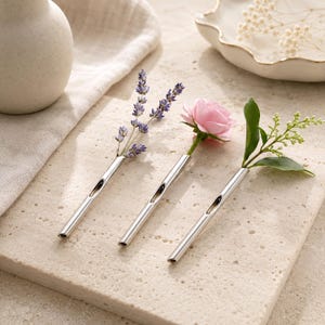 Könnte beinhalten: Drei schlanke, zylindrische Knospenvasen aus silberfarbenem Metall, die nebeneinander angeordnet sind. Jede Vase enthält ein einzelnes botanisches Element: einen Zweig lila Lavendel, eine zartrosa Rose und einen kleinen grünen Blattzweig mit Knospen.