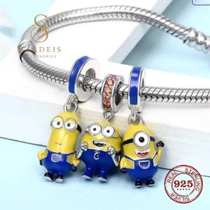 Puede incluir: Una pulsera de cadena de serpiente plateada con tres dijes colgantes con forma de personajes de dibujos animados amarillos y azules. Los dijes tienen detalles en esmalte y la pulsera incluye una cuenta espaciadora central con piedras naranjas.