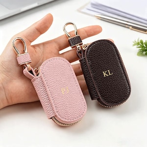 May include: Two oval-shaped key cases with a textured pebble grain finish, one in light pink and one in dark brown. Each case features a gold-toned zip, a matching metal swivel clasp, and gold-embossed initials reading PJ and KL respectively.