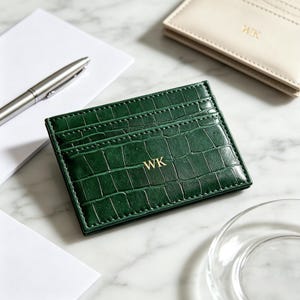 May include: A rectangular dark green card holder with a crocodile skin texture and gold embossed initials WK centered on the front. The card holder features multiple horizontal slots for cards and is positioned on a white marble surface.