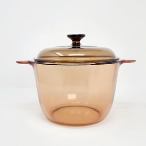 May include: A translucent amber-colored glass cooking pot with a matching lid. The pot features two integrated side handles and a central knob on the lid. The glass is smooth and clear, allowing light to pass through the warm, brownish-orange tint.