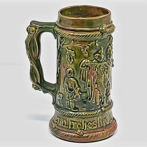 May include: A ceramic beer stein with a metallic green and bronze glaze. The exterior features embossed figures of people in a forest setting, a rope-like border at the top and bottom, and a handle shaped like twisted branches.