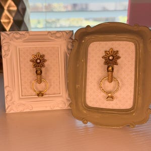May include: Two small rectangular frames, one white and one beige, each containing a white background with a subtle cross pattern. Centered in each frame is a gold-toned flower ornament with a hanging gold ring holding a clear gemstone.