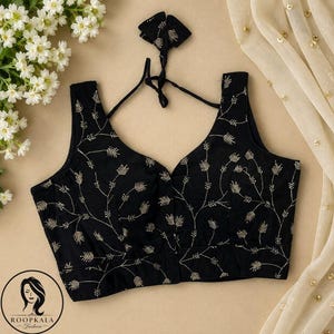 Puede incluir: Una blusa negra sin mangas con un escote en V profundo y un delicado patrón de enredaderas bordado en hilo dorado. La prenda incluye tirantes negros finos para atar en la espalda y un diseño de corpiño estructurado con una banda ancha.