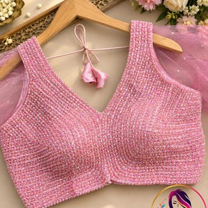 May include: A sleeveless pink blouse featuring a deep V-neckline and a dense, all-over embellishment of iridescent beads and sequins. The garment includes sheer pink tulle sleeves and a delicate tie-back closure with fabric tassels at the nape.