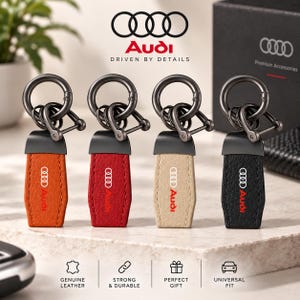 May include: Four keychains featuring orange, red, beige, and black leather straps with the Audi logo. Each keychain has a dark metal hardware attachment with a circular ring and a shackle-style connector. The text Audi Driven By Details is visible.