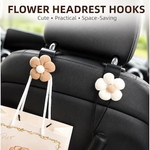 May include: Two black car headrest hooks featuring beige and white corduroy flower accents. The text FLOWER HEADREST HOOKS Cute • Practical • Space-Saving is visible at the top. The hooks are attached to a black car seat, holding a shopping bag.