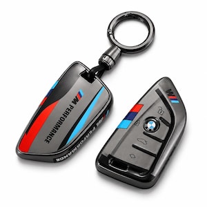 Op de afbeelding: Een donkergrijze metalen autosleutelhouder met het M Performance-logo en kenmerkende blauwe, paarse en rode strepen. De set bevat een bijpassende sleutelcover en een ronde metalen sleutelhanger, afgewerkt met een strakke, glanzende laag.
