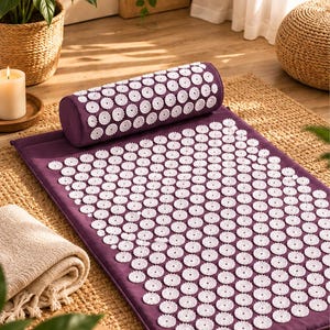 May include: A deep purple acupressure mat and a matching cylindrical bolster pillow, both featuring numerous white plastic circular discs with small, sharp spikes arranged in a uniform grid pattern across the entire surface of the fabric.