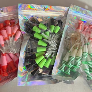 May include: Three holographic resealable pouches containing strings of miniature lava lamp-shaped lights. The first bag holds red and pink lamps, the second contains black and neon green lamps, and the third features white lamps with red or green stripes.