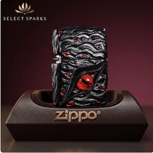 Può includere: Un accendino Zippo rettangolare con una superficie nera testurizzata, motivi a occhio rosso incastonati e un occhio rosso centrale prominente. L'accendino è esposto su un supporto marrone con il logo Zippo e il testo Select Sparks in alto.