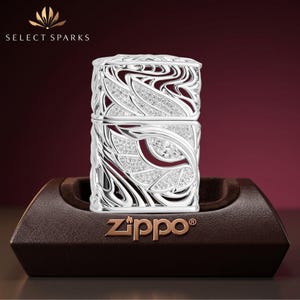 Puede incluir: Un encendedor Zippo plateado con un intrincado diseño de filigrana calada con patrones de remolinos y cristales transparentes incrustados. El encendedor se exhibe en un soporte contorneado marrón oscuro con el logo Zippo en la base.