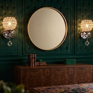 May include: A round mirror with a thin gold frame centered between two wall sconces. Each sconce features a spherical shade composed of clear crystal beads and a single dangling crystal teardrop, emitting a warm light from the center.
