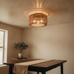 May include: A cylindrical ceiling light fixture featuring a woven tan rattan shade. The light is illuminated, casting a distinct radial pattern of light and shadow across the ceiling surface. The structure is circular with a lattice design.