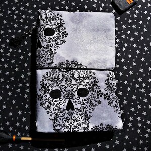May include: Two gray and black skull-themed pouches stacked on a black star-patterned surface. The skulls are created with intricate black floral designs. A black tassel hangs from the top pouch. The background is a black fabric with white stars.