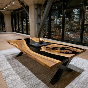 Pode incluir: Uma mesa de jantar de madeira com borda natural, apresentando um detalhe central em resina preta e um padrão de veios de madeira natural. A mesa é sustentada por uma base de metal preto em forma de X e contém uma bandeja quadrada de pedra.