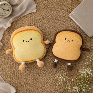 May include: Two plush toys shaped like slices of bread. The left slice is light yellow with a smiling face, while the right slice is darker brown with a sad face. Both plushies feature small arms and legs attached to their soft, textured bodies.