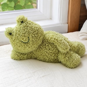 Op de afbeelding: Een lichtgroene pluche kikkerknuffel met een textuur van ruige stof. Het knuffeldier heeft een ronde kop met twee kleine uitstekende ogen, een geborduurde donkergroene gebogen mond en een zacht lichaam in een liggende houding.