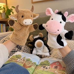 Puede incluir: Dos marionetas de peluche, una de color canela con cuernos y otra blanca con manchas negras y hocico rosa, se sostienen en alto. Un tercer juguete de peluche de vaca en blanco y negro se sitúa en el centro sobre un libro infantil ilustrado.