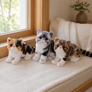 Puede incluir: Tres juguetes de peluche con forma de gato, cara plana y grandes ojos oscuros. El gato de la izquierda tiene manchas tricolores naranja, negras y blancas. El del medio es gris y blanco con rayas. El de la derecha es marrón y negro.