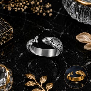 May include: A silver-toned open ring featuring a fish design where the head and tail meet at the opening. The metal surface has a textured, spotted pattern across the fish body. A smaller circular inset shows the same ring in a gold-toned finish.