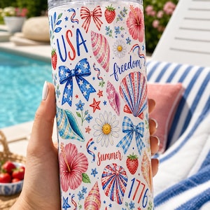 May include: A clear cylindrical tumbler with a metal straw featuring a white background decorated with red, white, and blue patterns including seashells, hibiscus flowers, strawberries, daisies, bows, and the text USA, freedom, summer, and 1776.