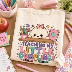May include: A cream-colored sweatshirt featuring a graphic of a smiling dumpling character with a pink bow, a pencil cup, and a notebook. The text reads TEACHING MY LITTLE DUMPLINGS, accompanied by colorful illustrations of stars, hearts, and crayons.