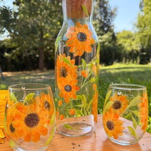 Peut inclure: Une carafe en verre transparent avec un bouchon en liège et deux verres sans pied assortis, tous peints à la main avec des tournesols orange et jaunes vifs, des feuilles vertes et de petites abeilles. L'ensemble présente un motif floral.