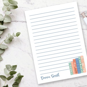 May include: A white lined notepad with a blue line pattern. The bottom right corner features a stack of colorful books with floral designs. The notepad is personalized with the name "Dorina Smith".